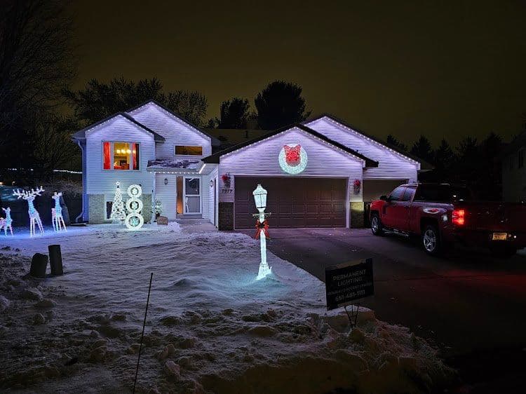 Christmas lights decorate a home with snowy yard, reindeer figures, and a festive red truck.