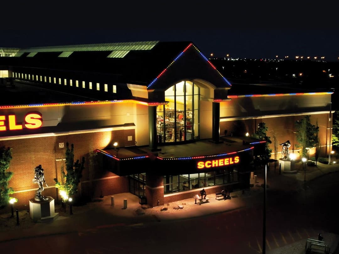 Scheels store illuminated at night with colorful lights and outdoor sculptures.