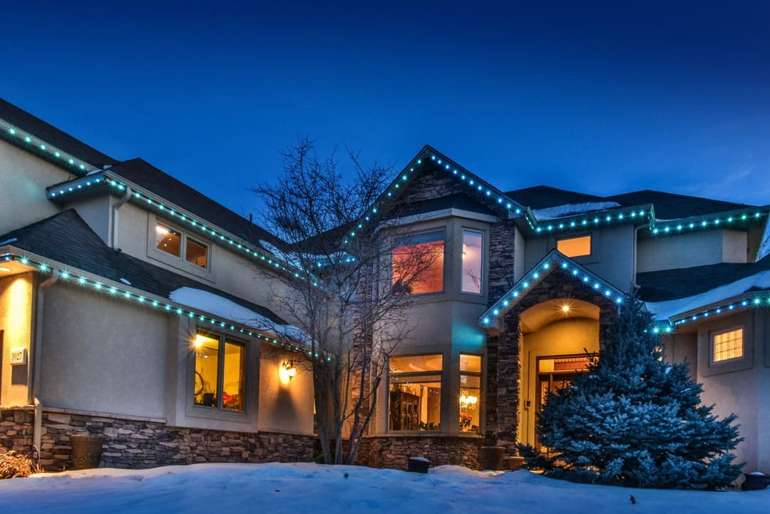 Twilight view of a beautifully lit home adorned with blue holiday lights and snow-covered landscaping.