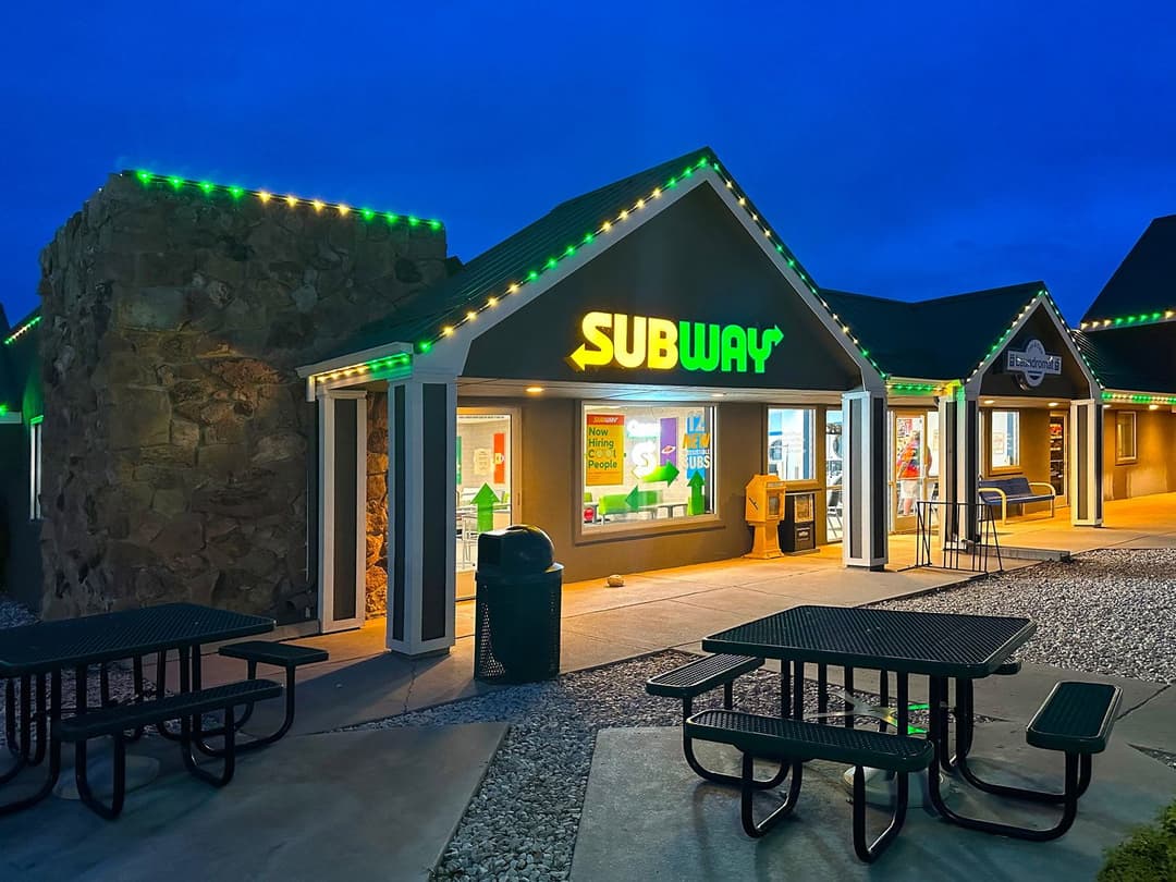 Subway restaurant exterior at dusk with bright green and yellow signage and outdoor seating.