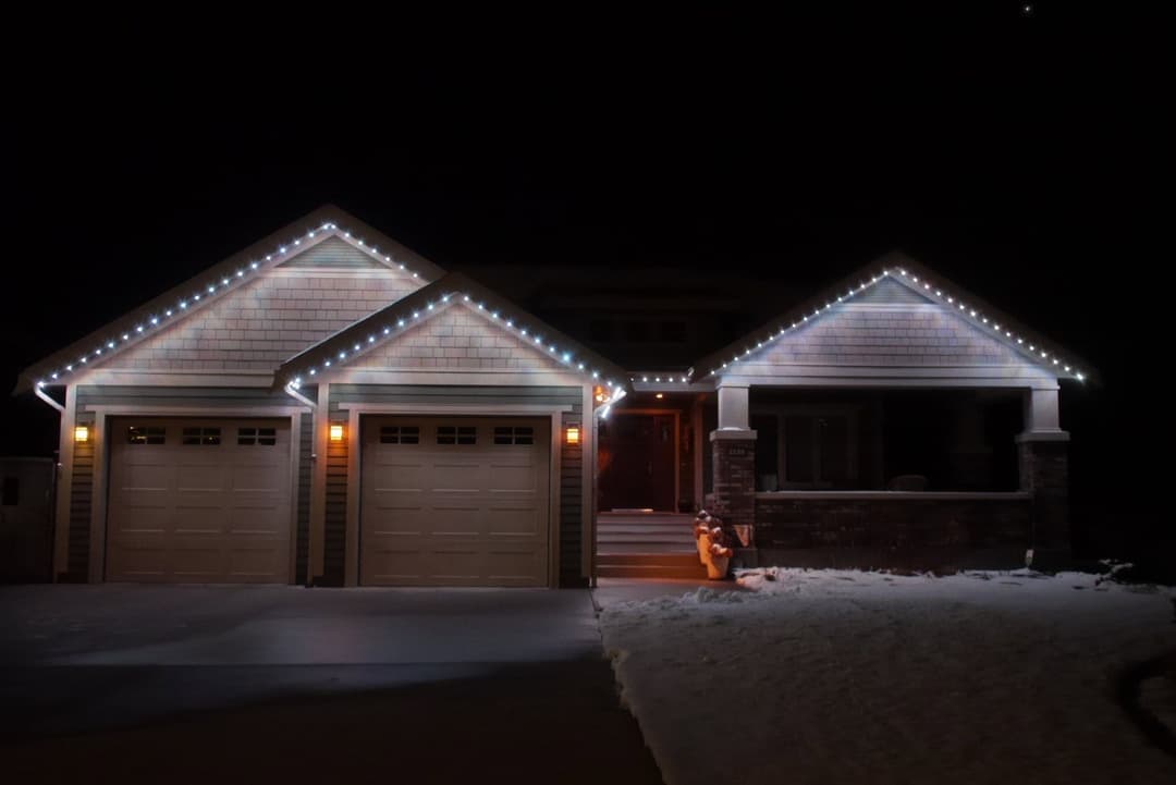Decorated house at night with holiday lights on eaves and snow-covered driveway.