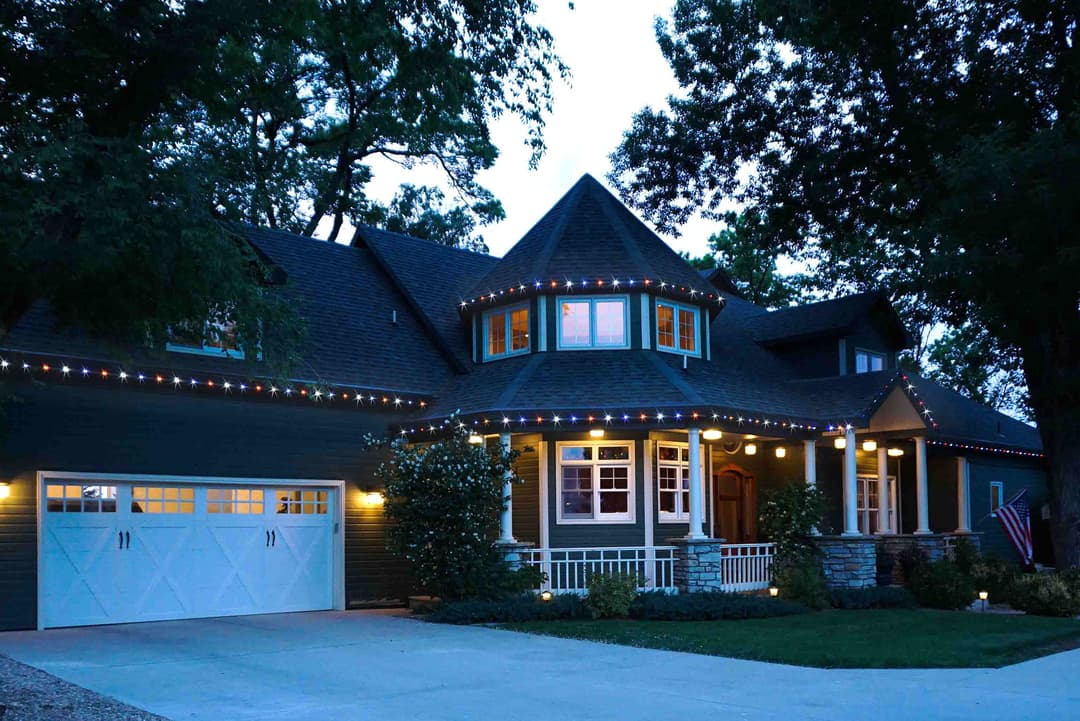 Elegant house with festive lights, large porch, and landscaped yard at dusk.