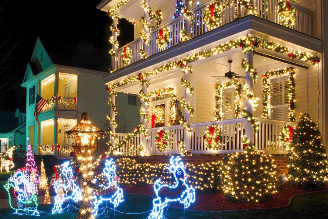 Festively decorated house with Christmas lights, wreaths, and illuminated reindeer in yard.